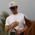 Man wearing an Alpine Days Rodeo cap and holding a lasso, standing next to a horse with a blurred background