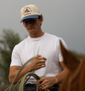 Man wearing an Alpine Days Rodeo cap and holding a lasso, standing next to a horse with a blurred background