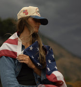 Person wrapped in an American flag with a raspberry days cap, standing outdoors against a dark sky.